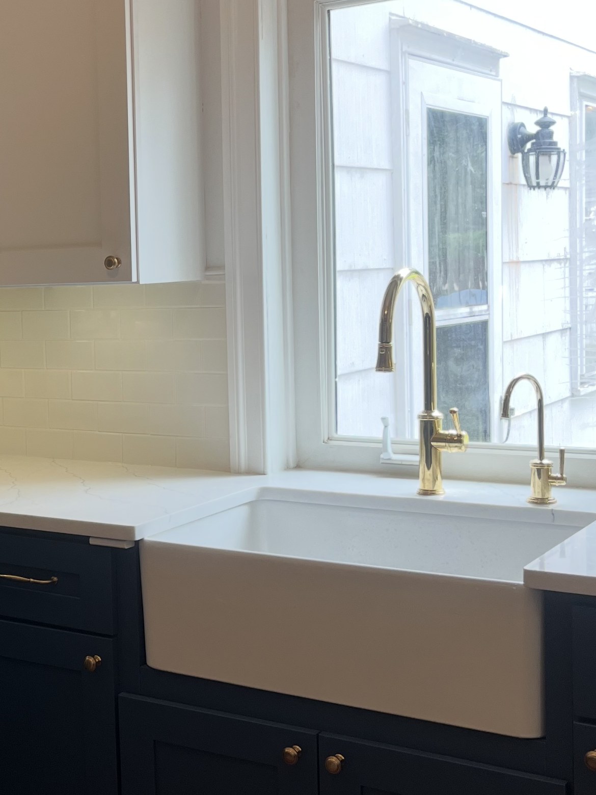 Farmhouse sink under the kitchen window, brass faucet, white counter running out to the sill, navy cabinets below
